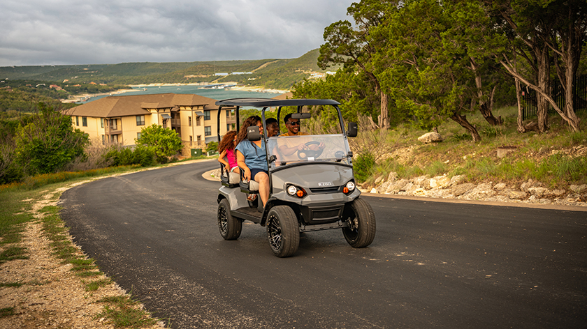 A group of friends drive an E-Z-GO golf cart up a hill with the horizon visible in the background.