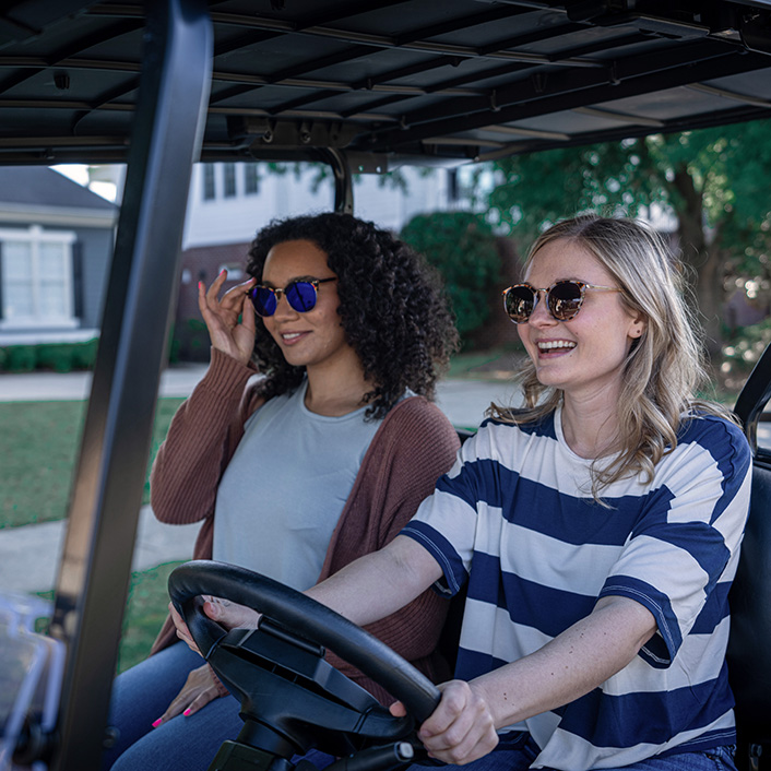 Two women wearing sunglasses smile while driving an E-Z-GO golf cart around a neighborhood.
