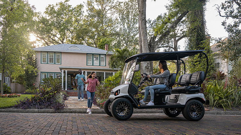 A child and father walk toward a woman driving an E-Z-GO golf cart.