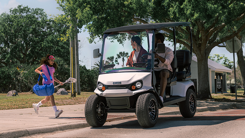 Two adults pick up a child carrying a bag in their E-Z-GO golf cart.