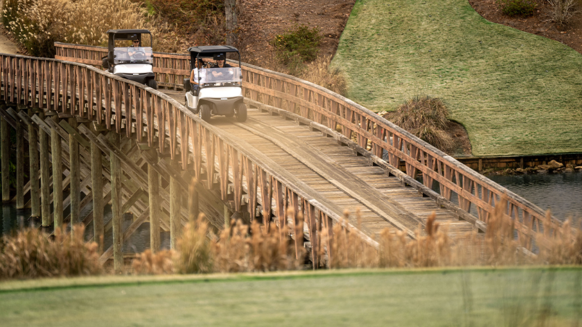 Two E-Z-GO golf carts drive over a bridge on a golf course.