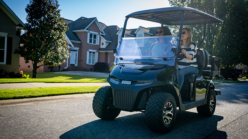 Two women drive an E-Z-GO golf cart on a neighborhood road.