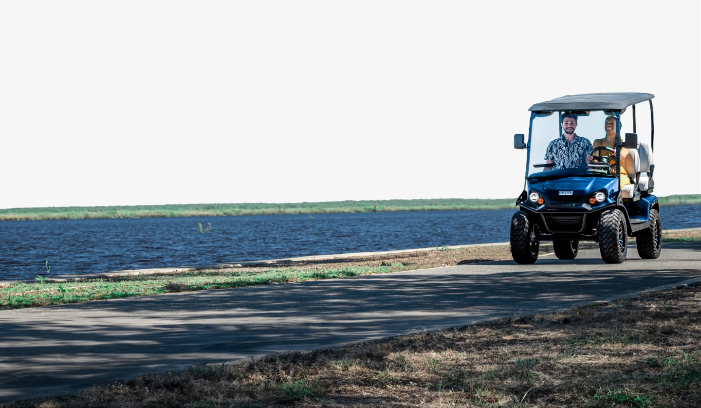 A driver and passenger ride an E-Z-GO golf cart alongside a body of water.
