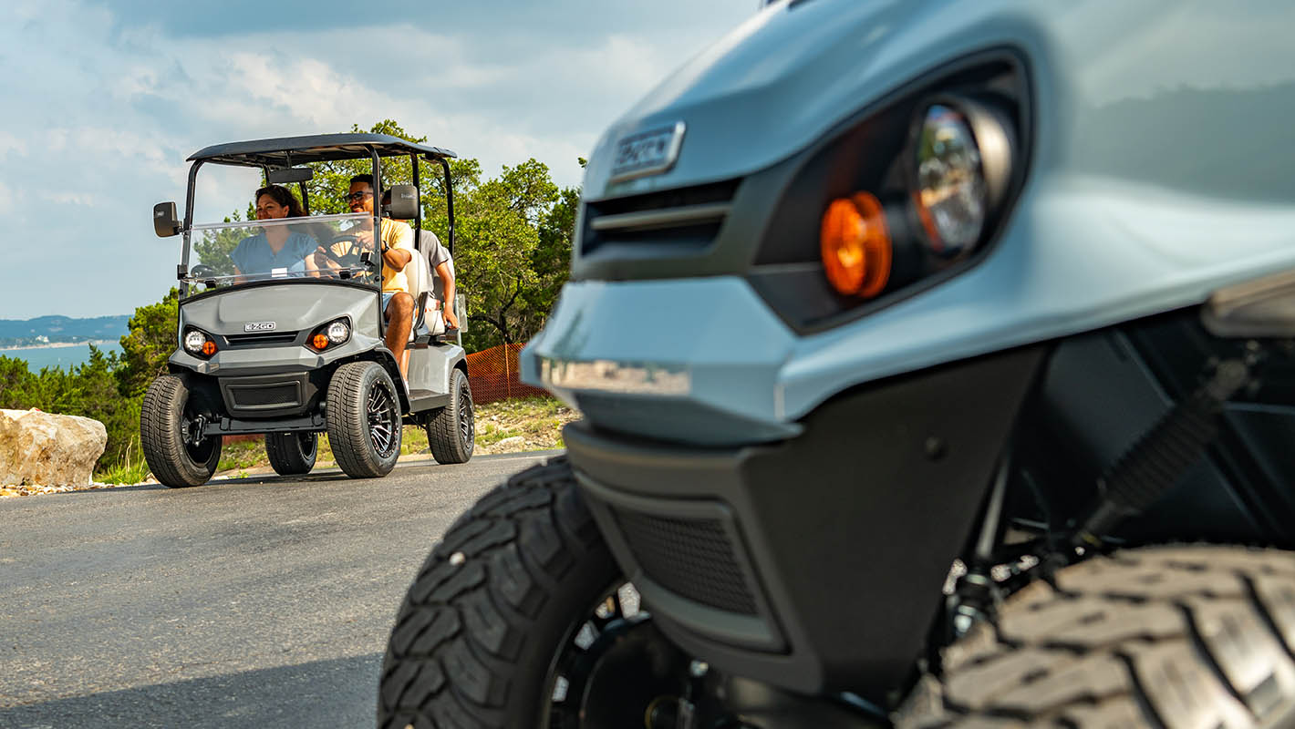 An E-Z-GO golf cart being driven down a road, another E-Z-GO frontend in the foreground.
