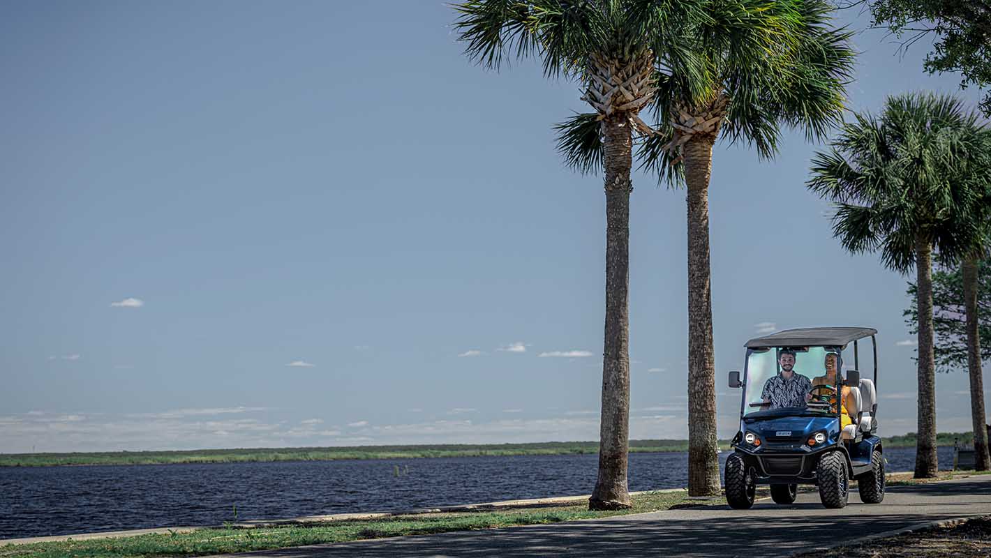 An E-Z-GO golf cart driving along a path next to a body of water lined with palm trees.